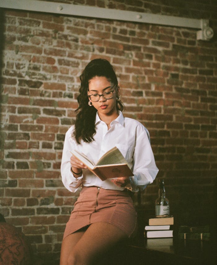 classes-03 A young woman in a casual outfit reads a book indoors against a brick wall.